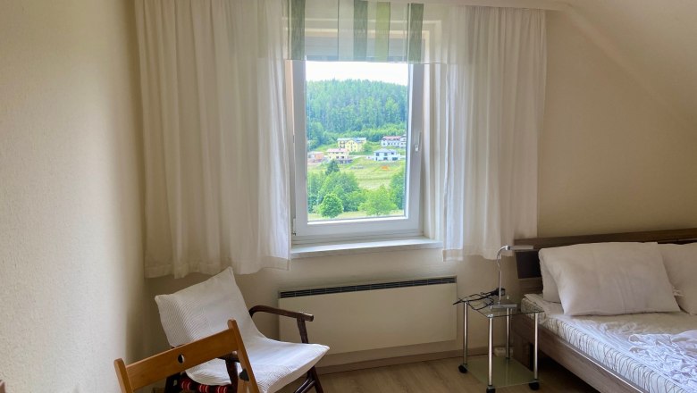 Bedroom2(c)wieneralpen, © Wiener Alpen Bright bedroom with window view of green landscape, bed, chair and table.