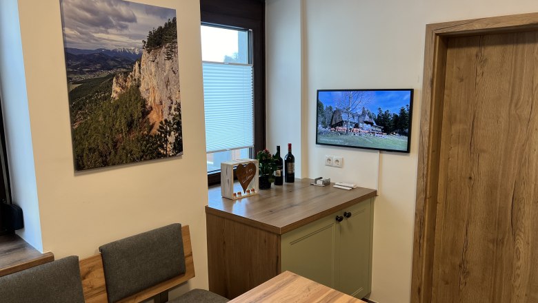 Kitchen, © Anton Sommer Kitchen with wooden furniture, a landscape painting on the wall and a TV showing a picture of a house.