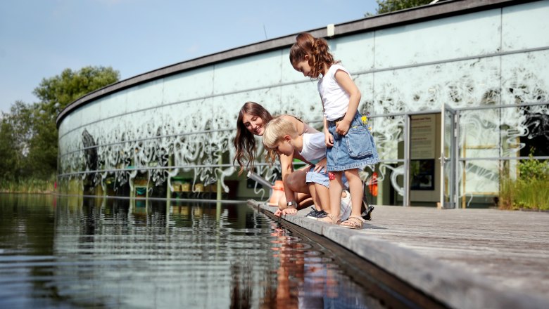 UnterWasserReich Schrems_WF244631 © Waldviertel Tourismus, weinfranz, © Waldviertel Tourismus/weinfranz Three people looking at water on a jetty in front of a modern building.