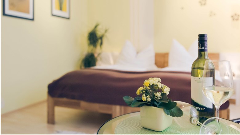 Apartment & guest room "Vino Verde", © Fam. Willmann A cozy bedroom with a bed in the background and a table with a bottle of wine and a glass in the foreground.