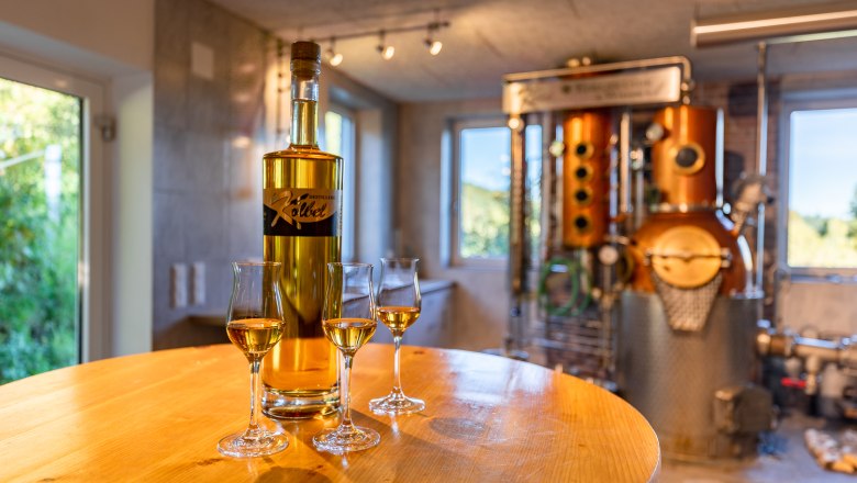 Kölbel family firing plant, © Wiener Alpen, Christian Kremsl A bottle and three half-filled glasses stand on a round wooden table in front of a still.