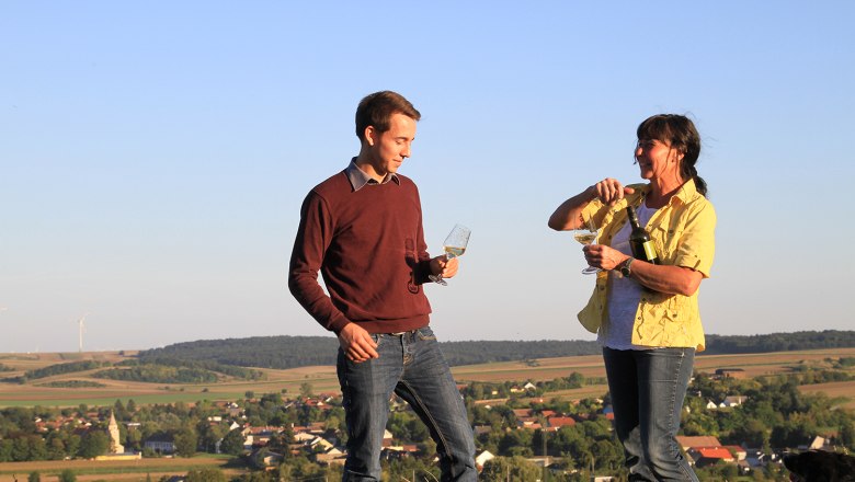 Winery Thüringer, © Weingut Thüringer Two people are standing on a hill with wine glasses in their hands, in the background a rural landscape with wind turbines.