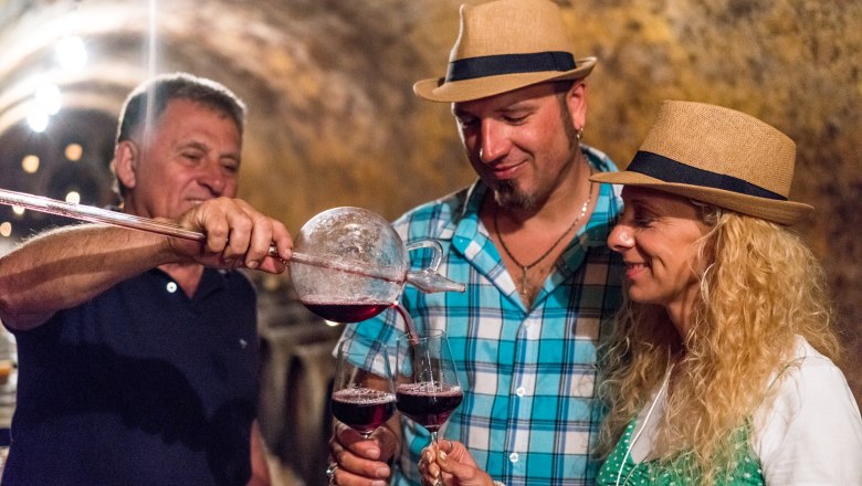 Wine tasting, © Fam. Zesch Three people in a wine cellar, two wearing straw hats. A man is pouring red wine into glasses.