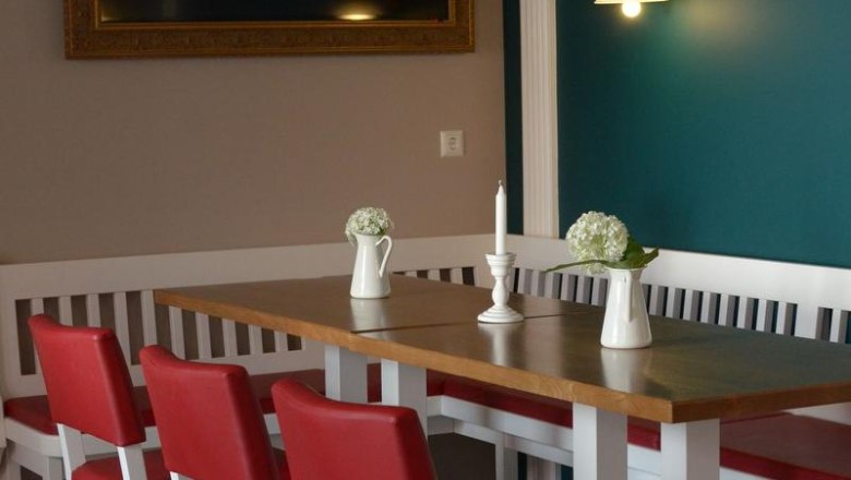 Chamber and paddock, © Julia Pfeiffer / Fam. Engelbrecht A dining area with red chairs, a wooden table, white vases and a candle. On the wall, a large, empty picture frame and a lamp.