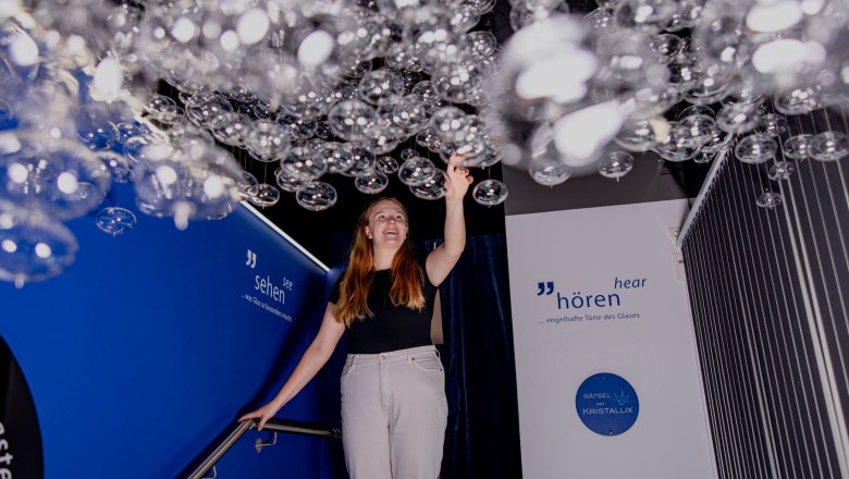 KRISTALLIUM glass.experience., © Waldviertel Tourismus, Matthias Streibel A woman touches hanging glass bubbles in an exhibition with blue walls and inscriptions.
