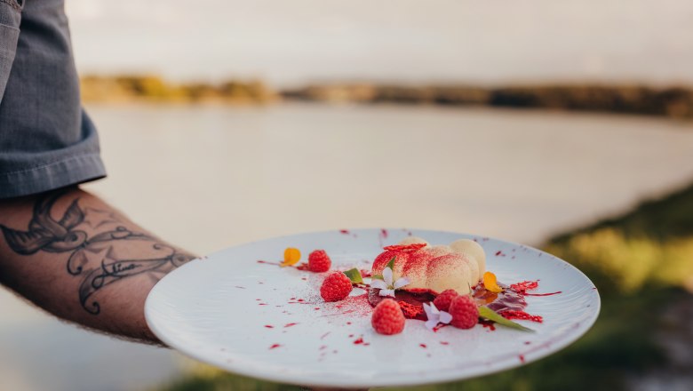 Cloud 7: White chocolate mousse with raspberry jelly & raspberry dust, © Niederösterreich Werbung/Daniela Führer A plate of white chocolate mousse, raspberries and flowers, held by a tattooed hand in front of a lake.