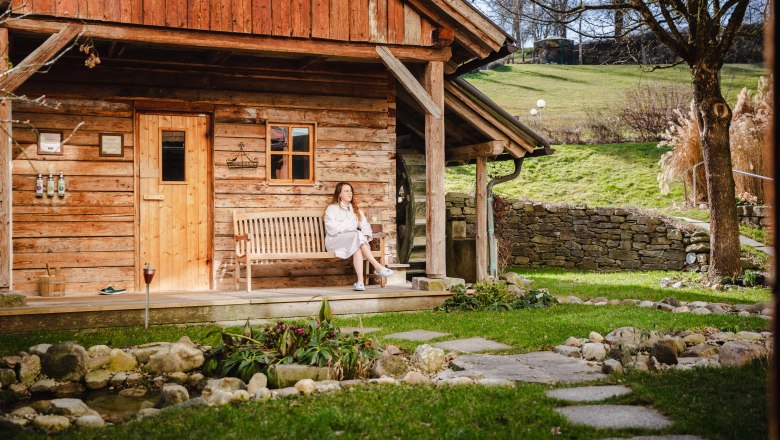 RelaxResort Kothmühle, © Johanna Meinschad Woman in bathrobe sitting on a bench in front of a wooden hut in the countryside.