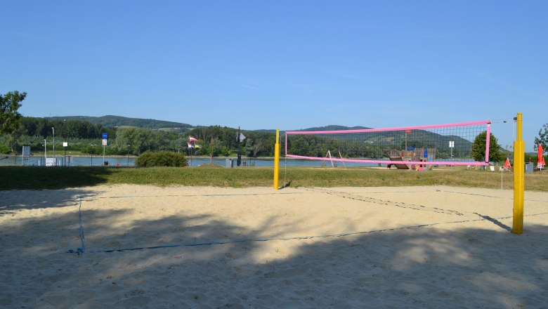 Beach volleyball court, © Stadtgemeinde Korneuburg An empty beach volleyball court with yellow posts and a pink net in front of a river and green hills in the background.