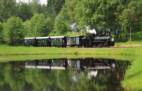 Waldviertel Railway, © knipserl.at A historic steam locomotive pulls green wagons through a verdant landscape reflected in a tranquil pond.