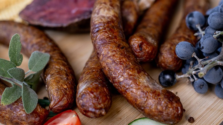 At the Arkadenhof, products from our own production, © Martina Siebenhandl Close-up of sausages, grapes, tomatoes, cucumbers and onions on a wooden board.