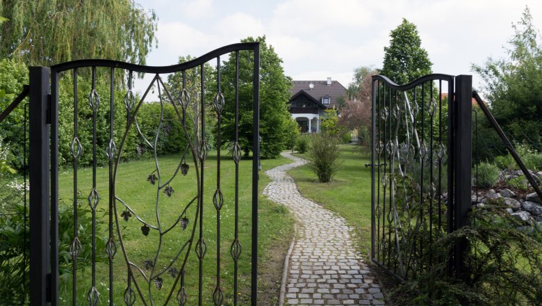 Gate to the garden, © Leopold Pluschkowitz An open wrought-iron gate leads to a paved path that leads through a green garden to a house in the background.