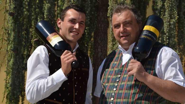 Waldschütz family, © Familie Waldschütz Two men in traditional dress hold large bottles of wine in front of a tree.