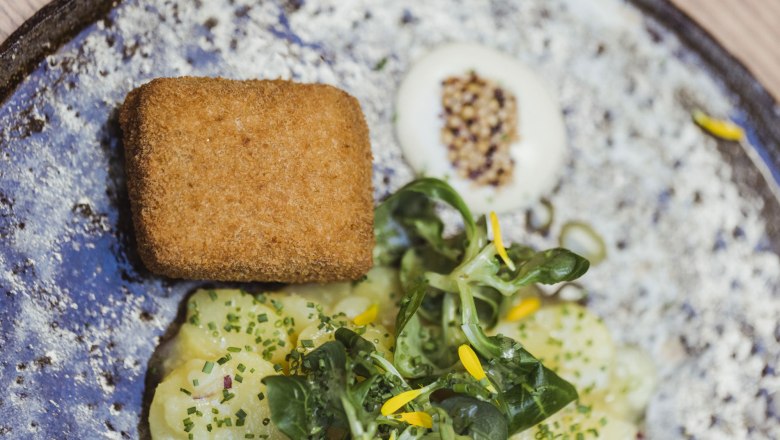 Veal head on a bird's head and apple salad, © Niederösterreich Werbung/David Schreiber A plate of breaded veal head, potato salad and lamb's lettuce, garnished with yellow flowers.