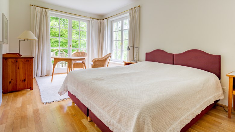 Bedroom, © Wiener Alpen / Christian Kremsl Bright bedroom with large bed, wooden floor, two wicker chairs and table in front of large windows with a view of the greenery.