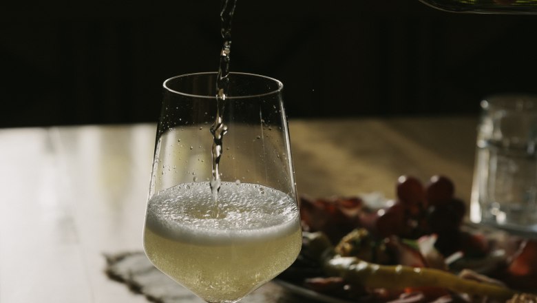 Pouring wine, © Weinviertel Tourismus / Sophie Menegaldo A glass of white wine is poured on a table, with a plate of snacks in the background.
