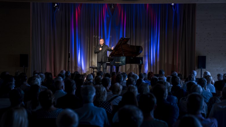 Cultural Summer Semmering Event, © wieneralpen_herbst A man stands on a stage with a grand piano and speaks into a microphone in front of an audience.