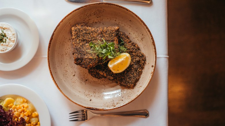 Waldviertel carp fillet from Zwettl Abbey breaded in gray poppy seeds, © Niederösterreich Werbung/Daniela Führer Grey poppy seed breaded carp fillet with lemon slice and herbs on a plate, side dishes next to it.