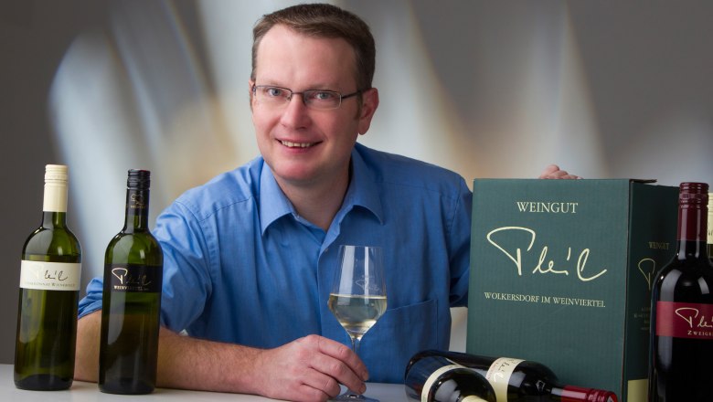 Christian Pleil, © Fotostudio Semrad A man in a blue shirt holds a wine glass and sits in front of bottles of wine and a wine crate from the Pleil winery.