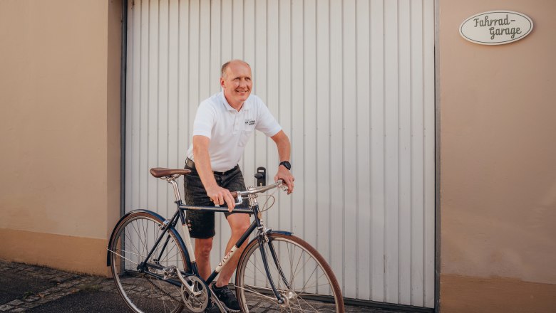 Bike garage, service, rental, tours, © Niederösterreich Werbung/Daniela Führer Man in white polo shirt rides bicycle in front of garage door.