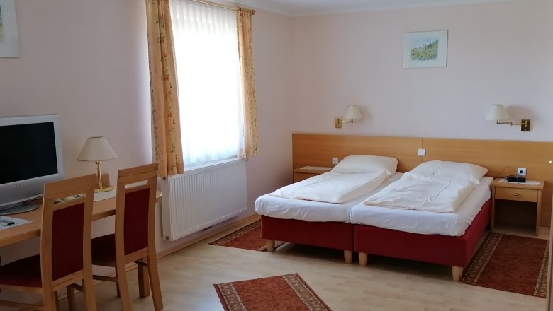 Double room, © Wiener Alpen A double room with two single beds, a desk with chairs, a TV and a window with curtains.