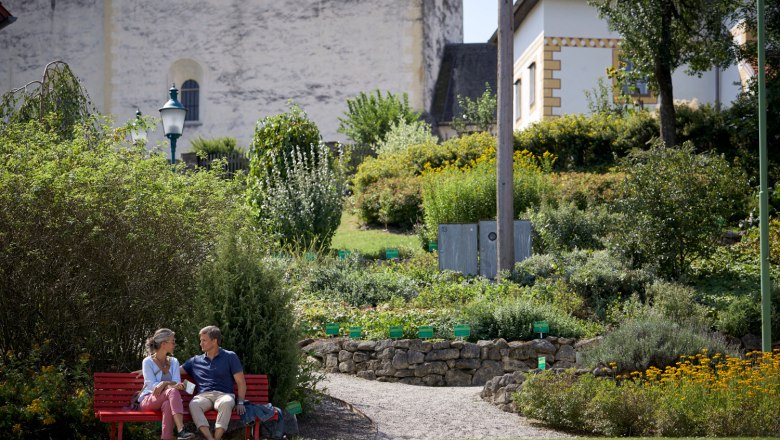 In front of the fortified church in Bad Schönau, © Wiener Alpen, Florian Lierzer A couple sits on a red bench in a garden in front of the fortified church in Bad Schönau.