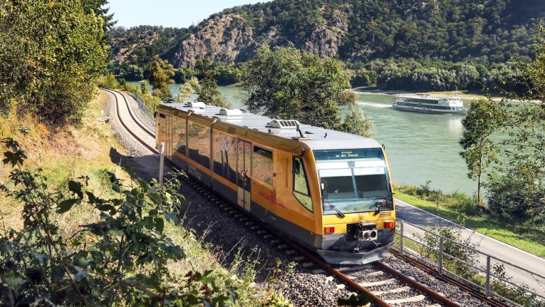 Wachau Railway, © NÖVOG, weinfranz A yellow train of the Wachau Railway runs along a river, with a cruise ship and wooded hills in the background.