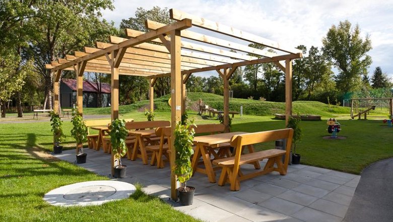 Arbor, © JUFA Wooden pergola with benches and tables outside, surrounded by lawn and trees.