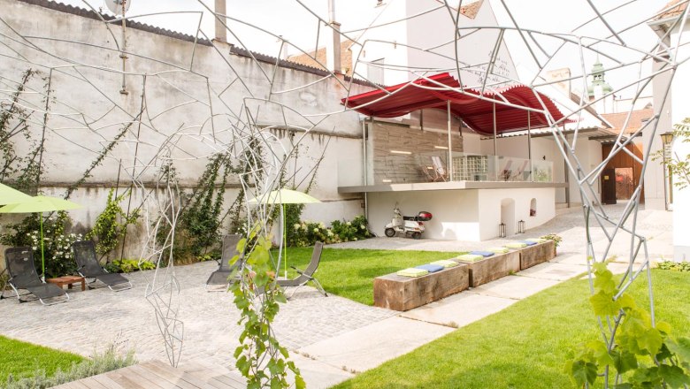 Klinger's guest house, © Astrid Bartl Modern courtyard with lawn, deckchairs, metal sculptures and a Vespa under a red canopy.