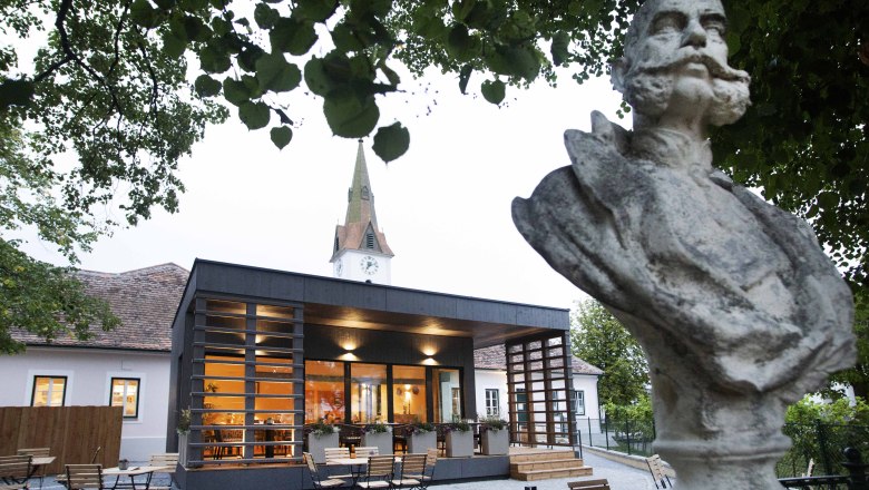 The FRANZ JOSEPH Wirtshaus on Kaiserplatz, © Astrid Bartl Bust in front of a modern building with a church tower in the background, surrounded by trees.