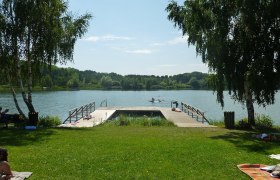 Langau mining lake, © Gemeinde Langau A jetty at Bergwerksee Langau with people swimming and sunbathing. Surrounded by trees and green meadows.