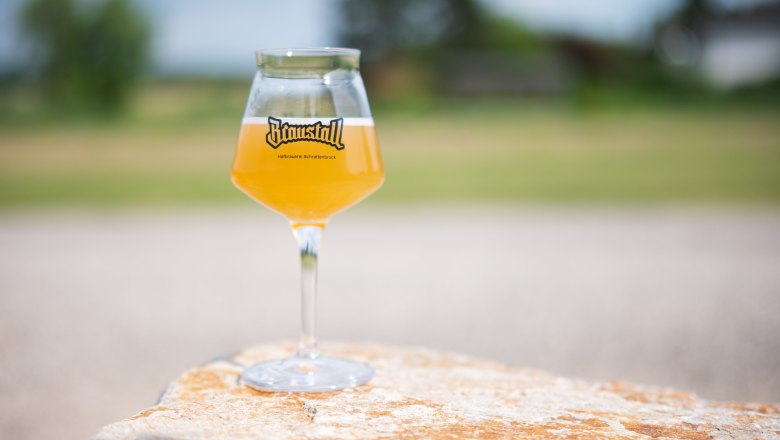 braustall_brewday, © Hofbrauerei Braustall_Georg Schwameis A glass of pale beer with the Braustall logo stands on a stone outside.