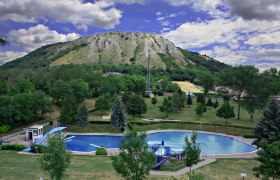 Mountain bath Hainburg/Danube, © Stadtgemeinde Hainburg/Donau An outdoor pool with a round pool and a slide, surrounded by trees, in front of a wooded hill with a rocky peak.