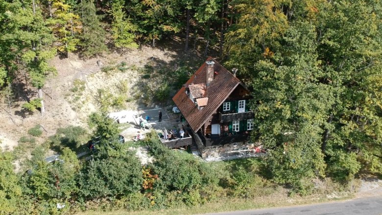 a9738793-a118-4c1a-a0a3-1f78cd2d9d26, © Martin Sturm Aerial view of a house in the forest with a red roof and green shutters, surrounded by trees.