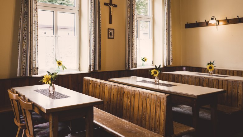 Where the village comes together, © Niederösterreich Werbung/David Schreiber A rustic room with wooden benches and tables, decorated with sunflowers in vases. A cross hangs on the wall.