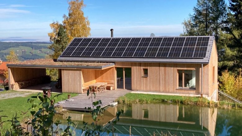 Vacation cottage Orthof, © Ferienhaus Gruber-List A modern wooden house with solar panels on the roof, surrounded by nature and a small pond in the foreground.