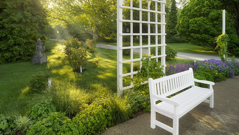 Baden Rosarium, © Rainer Mirau White bench in the rose garden with trellis and plants in the sunlight.