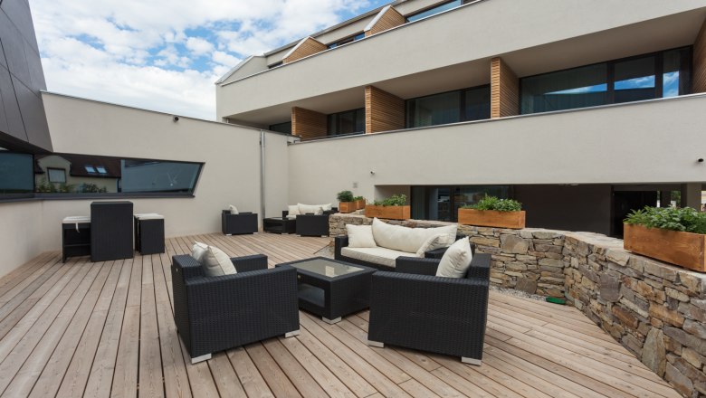 Terrace with lounge furniture, © KASTNER NEW MEDIA - kastner.tv Modern terrace with wooden floor, black rattan furniture and stone wall, surrounded by a modern building.