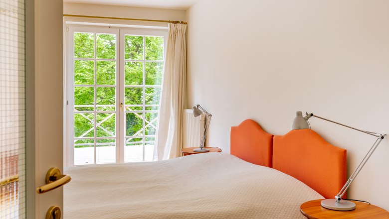 Bedroom, © Wiener Alpen / Christian Kremsl A bedroom with an orange headboard, a double bed and a window with a view of green trees.