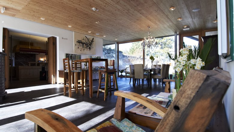 Breakfast room, © Hotel Landhaus Moserhof GmbH A modern breakfast room with wooden furniture, large windows and a chandelier. A wooden chair in the foreground and a dining table in the background.