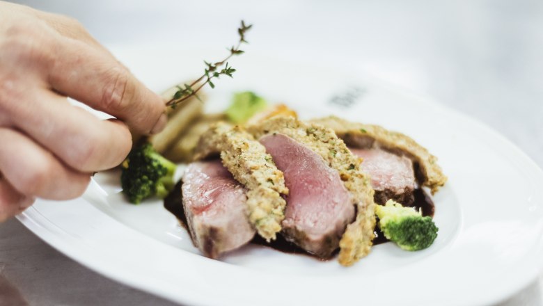 Pink saddle of venison with chestnut noodles, © Niederösterreich Werbung/David Schreiber Close-up of a pink roasted meat dish with herb crust, broccoli and a hand adding thyme.
