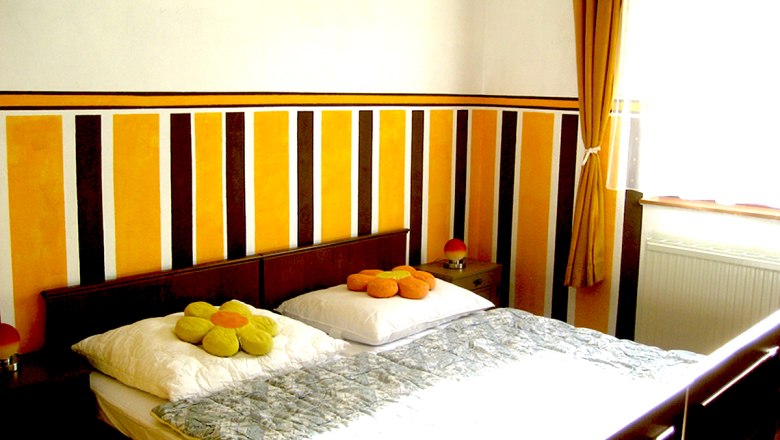 Bedroom 2, © Anke Spreitzer A bedroom with a double bed, striped wall and yellow curtains.