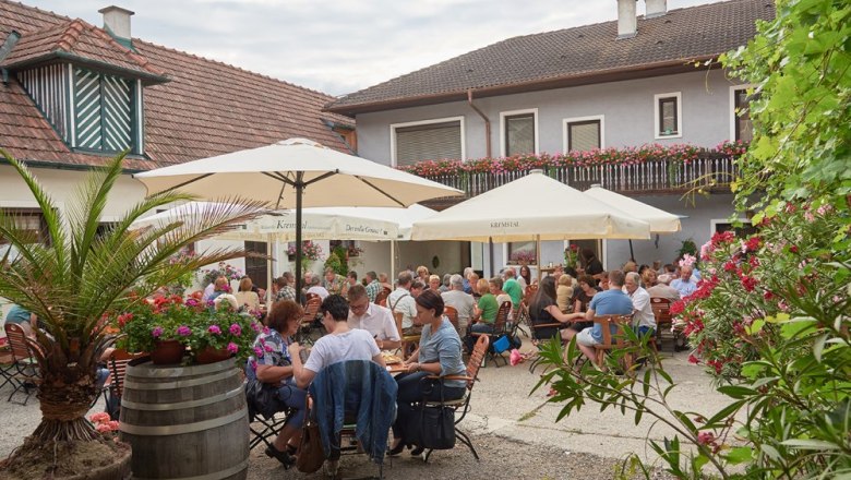 Courtyard wine tavern Weinhof Fink, © Weinhof Fink A lively courtyard of a wine tavern with lots of people at tables under umbrellas, surrounded by flowers and plants.