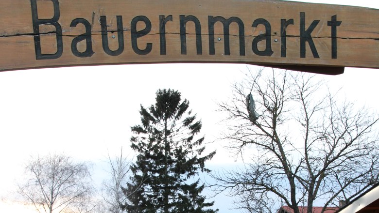 Simonsfeld farmers' market, © Josef Popp Entrance to a farmers' market with a wooden sign and a paved path surrounded by trees and buildings.