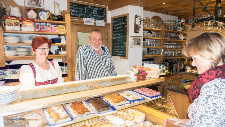 Waldhäusl, © Waldhäusl A man and a woman stand behind a counter in a café, while a customer stands in front. Various cakes are displayed on the counter.