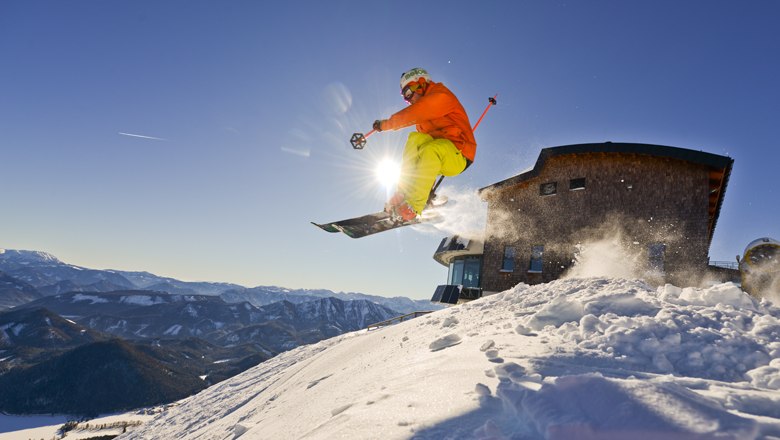 Action & fun on the Gemeindealpe-Terzerhaus, © Leiminger A skier jumps into the air in front of the Terzerhaus on the Gemeindealpe, surrounded by snow-covered mountains and a blue sky.