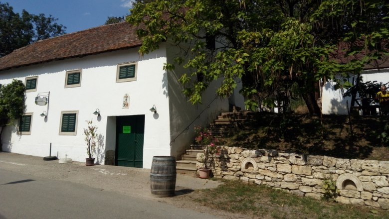 Himmelbauer Winery, © Weinstraße Weinviertel White building with green shutters and an open door, surrounded by trees and a wine barrel on the street.