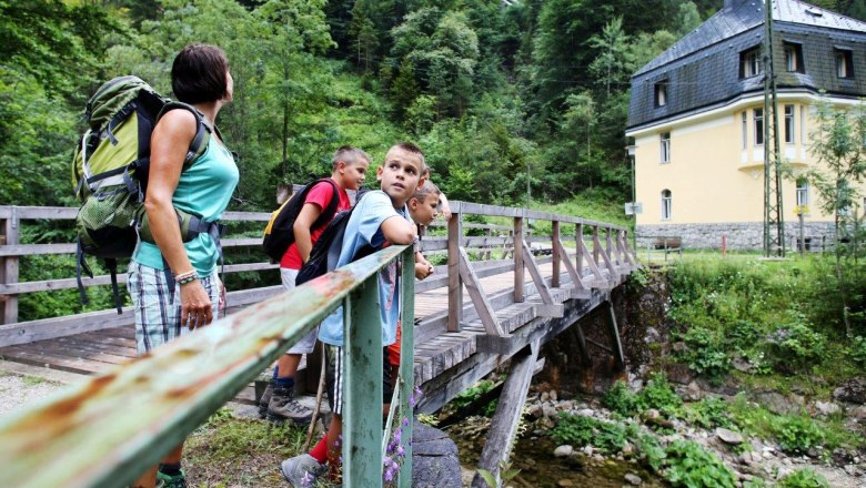 Power plant in Wienerbruck, © Mostviertel Tourismus, weinfranz.at A group of people are standing on a wooden bridge in a green, wooded setting. A yellow building can be seen in the background.
