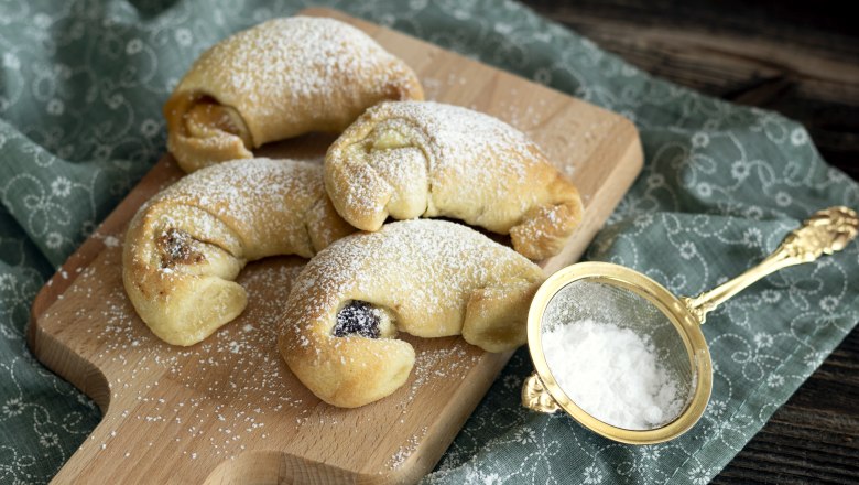 Four cream croissants on a wooden board, sprinkled with powdered sugar, next to a golden sieve on a patterned cloth., © Fam. Luckerbauer Four cream croissants on a wooden board sprinkled with powdered sugar, next to a sieve with powdered sugar.