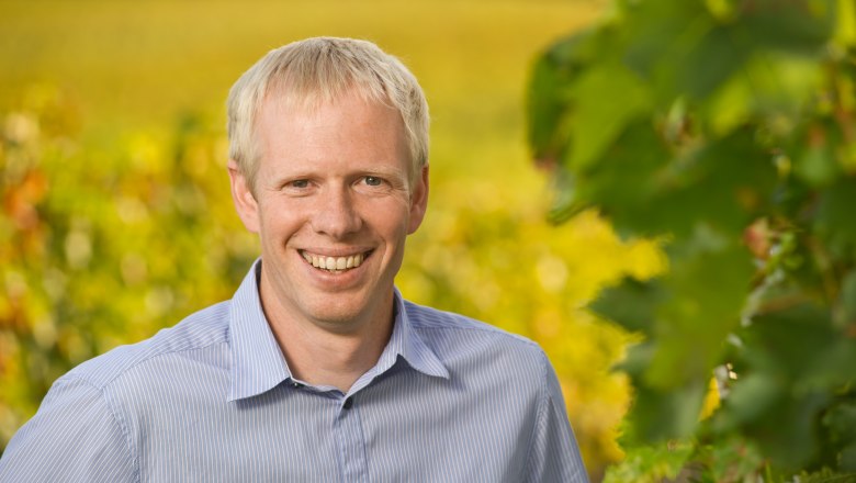 Winemaker Christoph Bauer, © Rita Newman A smiling man in a blue shirt stands in front of a blurred background with green leaves.