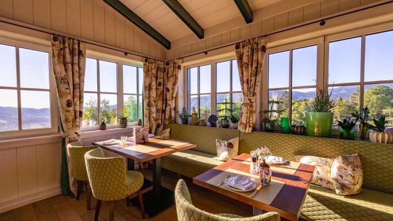 Hotel & Restaurant Knappenhof, © Niederösterreich Werbung / Maximilian Pawlikowsky Cozy dining area with large windows, green upholstered furniture and plants, with a view of the countryside.